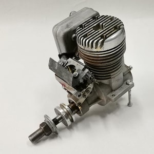 May include: A vintage model airplane engine, featuring a silver-colored cylinder head with cooling fins and a carburetor. The engine has a complex mechanical design with various components, including a fuel tank and a propeller shaft.