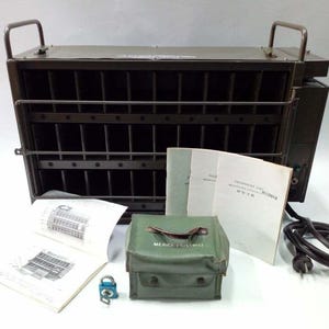 Peut inclure: Une bo&icirc;te m&eacute;tallique vert olive avec une fa&ccedil;ade grillag&eacute;e et deux poign&eacute;es, accompagn&eacute;e de manuels, d'une mallette verte avec l'inscription "MEDICE PROVISIO", d'un cadenas et d'un cordon d'alimentation. La bo&icirc;te a des barres verticales.