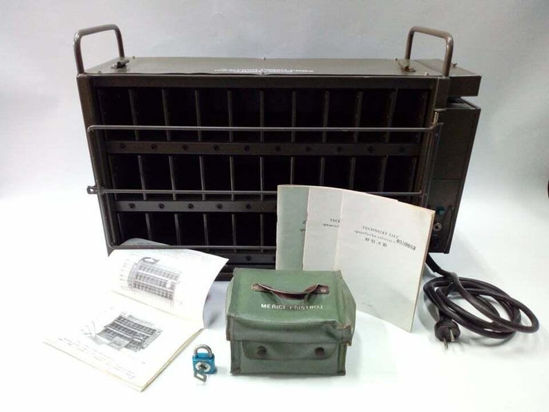 Peut inclure: Une bo&icirc;te m&eacute;tallique vert olive avec une fa&ccedil;ade grillag&eacute;e et deux poign&eacute;es, accompagn&eacute;e de manuels, d'une mallette verte avec l'inscription "MEDICE PROVISIO", d'un cadenas et d'un cordon d'alimentation. La bo&icirc;te a des barres verticales.