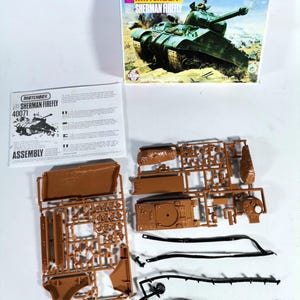 以下が含まれることがあります： マッチボックスシャーマンファイアフライの模型キット。箱には、戦闘シーンの緑色の戦車が描かれています。キットには、茶色のプラスチック部品、黒いトラック、説明書が含まれています。モデルは未組立です。
