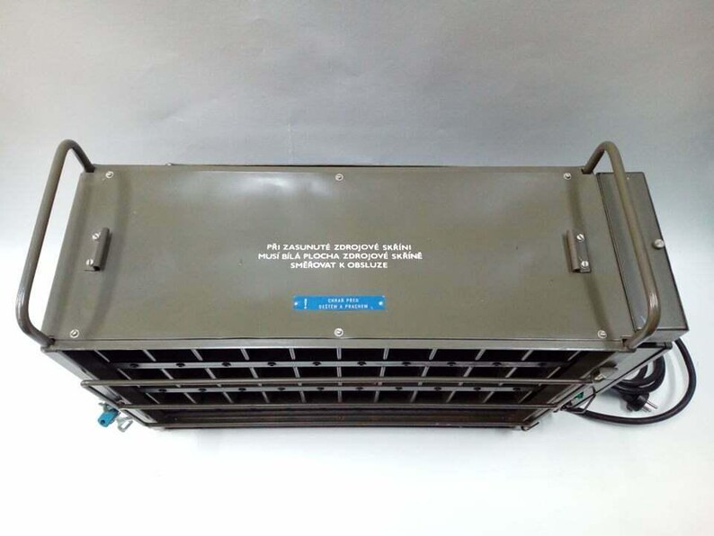 Peut inclure: Un appareil m&eacute;tallique vert olive de forme rectangulaire, avec une fa&ccedil;ade en grille et deux poign&eacute;es arqu&eacute;es. La surface sup&eacute;rieure comporte du texte et une &eacute;tiquette bleue. Un c&acirc;ble d'alimentation noir sort du c&ocirc;t&eacute; droit.