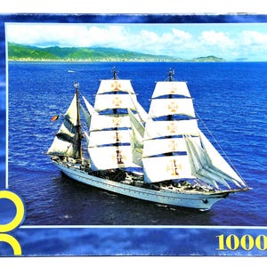 Op de afbeelding: Een puzzel van 1000 stukjes met een zeilschip op zee. Het schip heeft witte zeilen met gouden emblemen, tegen een blauwe zee en lucht. Het woord "Navigare" staat op de zijkant van de puzzel.