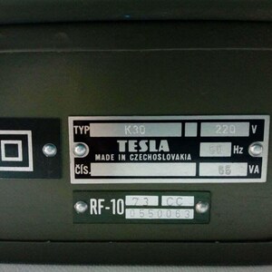 Peut inclure: Gros plan sur un bo&icirc;tier m&eacute;tallique vert fonc&eacute; avec plusieurs plaques attach&eacute;es. Les plaques affichent du texte, notamment "TESLA MADE IN CZECHOSLOVAKIA", "220 V" et "RF-10". Un symbole carr&eacute; est &eacute;galement visible.