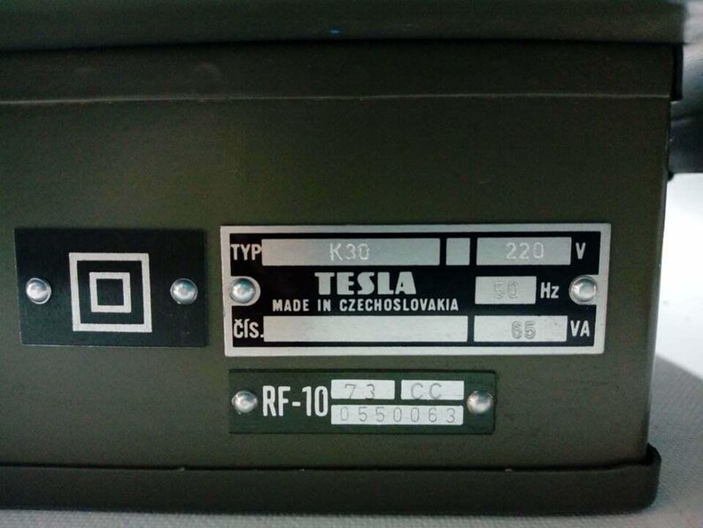 Peut inclure: Gros plan sur un bo&icirc;tier m&eacute;tallique vert fonc&eacute; avec plusieurs plaques attach&eacute;es. Les plaques affichent du texte, notamment "TESLA MADE IN CZECHOSLOVAKIA", "220 V" et "RF-10". Un symbole carr&eacute; est &eacute;galement visible.