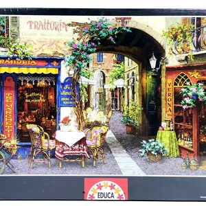Puede incluir: Un rompecabezas Educa de 1000 piezas con una ilustración detallada de una escena de calle europea. La imagen muestra una trattoria con asientos al aire libre, una entrada arqueada y tiendas con exhibiciones florales. La caja del rompecabezas es negra con el logotipo Educa Puzzle 1000.