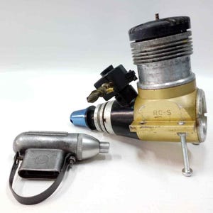 May include: A model airplane engine with a gold and silver finish, and a small silver starter. The engine has a black top and blue accents. The text "RC-S" is visible on the engine.