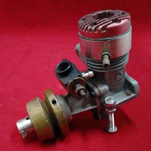 Può includere: Un motore modello vintage con una testa del cilindro rossa e un corpo argentato. Il motore ha componenti in ottone e argento, tra cui un volano e hardware di montaggio. Lo sfondo è rosso pieno.