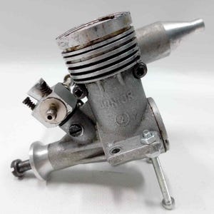 May include: A small, silver-colored Junior model engine with a cylindrical body and cooling fins. The engine has various mechanical components, including a carburetor, spark plug, and exhaust port. The word "JUNIOR" is visible on the engine's body.