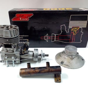 May include: A disassembled model engine with a silver cylinder head, a bronze exhaust pipe, and a silver carburetor. The engine is next to a black box with the text "T:gre" and "3000".