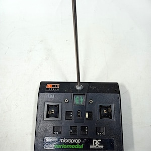 Peut inclure: Télécommande noire vintage avec une antenne argentée. La télécommande comporte plusieurs boutons, interrupteurs et cadrans. Le texte "microprop variomodul" et "BE Brand Elektronik" est visible à l'avant.