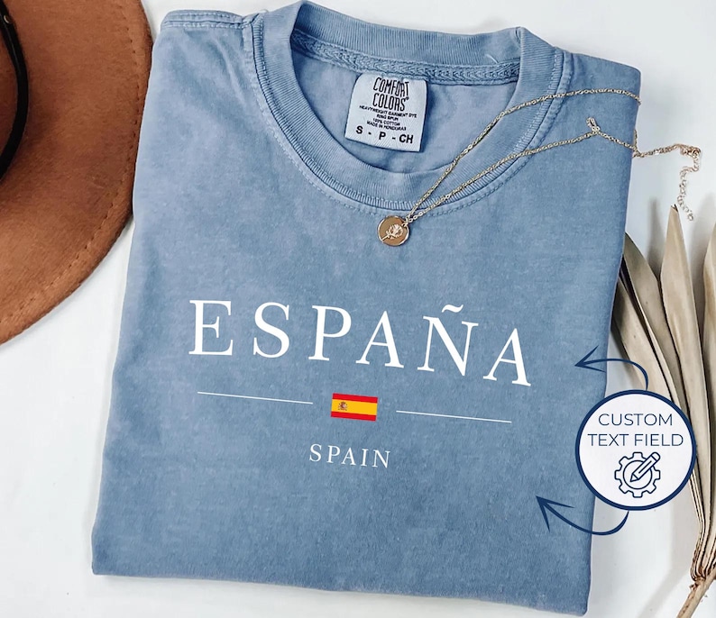 May include: A light blue t-shirt with the word "ESPA&Ntilde;A" in white, the Spanish flag, and the word "SPAIN" below. A gold necklace with a pendant rests on the shirt. The shirt has a Comfort Colors tag.