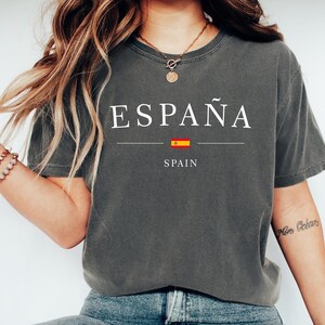 May include: Dark gray t-shirt with the word "ESPA&Ntilde;A" in white, a horizontal line, a small Spanish flag, and the word "SPAIN" in white. The shirt is a casual style.