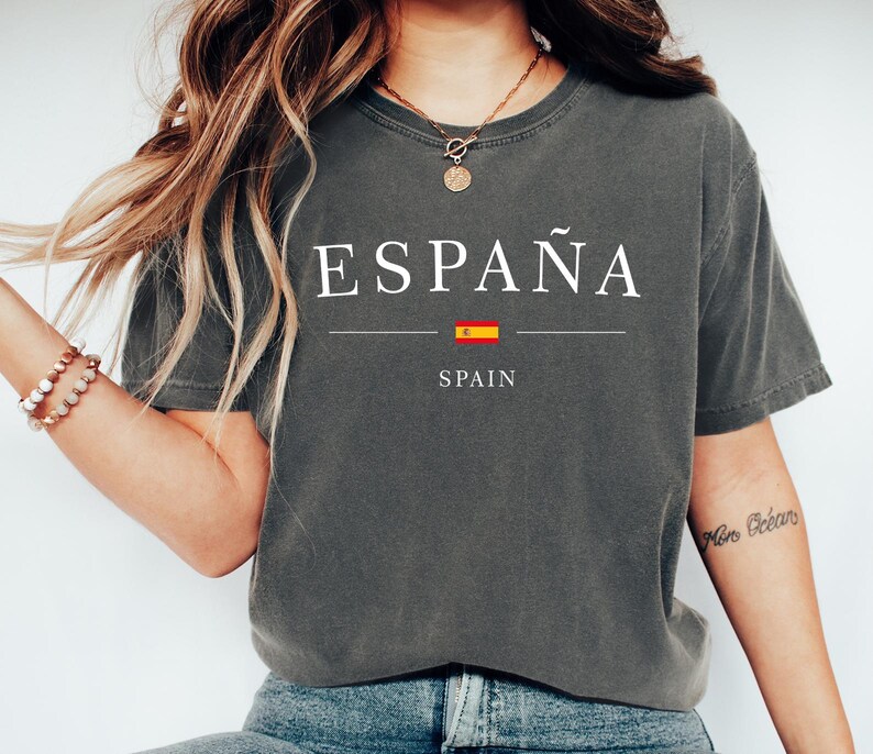 May include: Dark gray t-shirt with the word "ESPA&Ntilde;A" in white, a horizontal line, a small Spanish flag, and the word "SPAIN" in white. The shirt is a casual style.