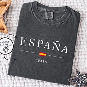 May include: Dark gray t-shirt with the word "ESPA&Ntilde;A" in white, a Spanish flag graphic, and the word "SPAIN" below. The shirt is made of a soft material and is laid flat on a white textured surface.