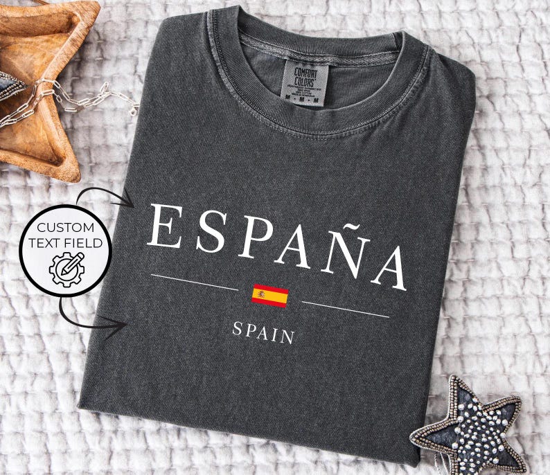 May include: Dark gray t-shirt with the word "ESPA&Ntilde;A" in white, a Spanish flag graphic, and the word "SPAIN" below. The shirt is made of a soft material and is laid flat on a white textured surface.