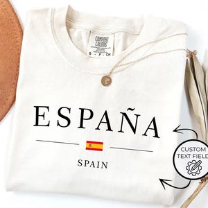 May include: Cream-colored t-shirt with the word "ESPA&Ntilde;A" in black, a Spanish flag graphic, and the word "SPAIN" underneath. A gold necklace and a brown hat are also visible.