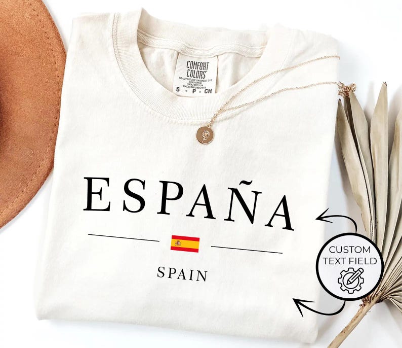May include: Cream-colored t-shirt with the word "ESPA&Ntilde;A" in black, a Spanish flag graphic, and the word "SPAIN" underneath. A gold necklace and a brown hat are also visible.
