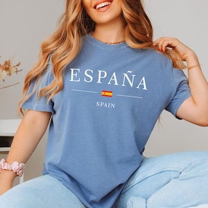 May include: A blue t-shirt with the word "ESPA&Ntilde;A" in white capital letters. Below the word is a horizontal line, a small Spanish flag, and the word "SPAIN". The shirt is made of a soft material.