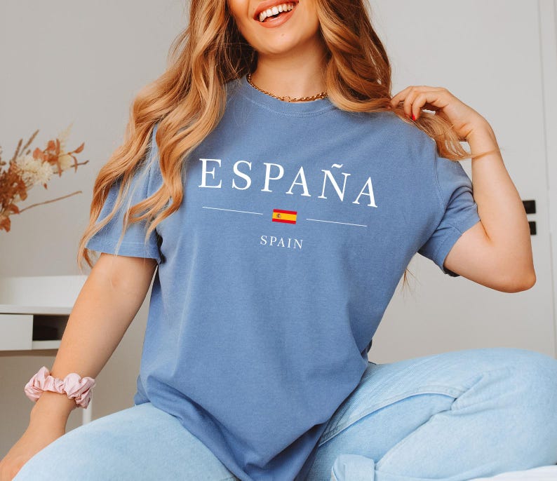 May include: A blue t-shirt with the word "ESPA&Ntilde;A" in white capital letters. Below the word is a horizontal line, a small Spanish flag, and the word "SPAIN". The shirt is made of a soft material.