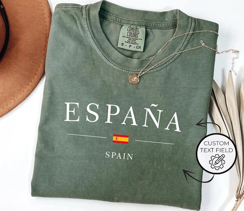 May include: An olive green t-shirt with the word "ESPA&Ntilde;A" printed in white, along with the Spanish flag and the word "SPAIN" below. A gold necklace with a pendant rests on the shirt. The shirt is folded.