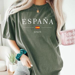 May include: Olive green t-shirt with the word "ESPA&Ntilde;A" printed in white, along with the Spanish flag and the word "SPAIN" underneath. The person is wearing a teal scrunchie and holding a pink mug.