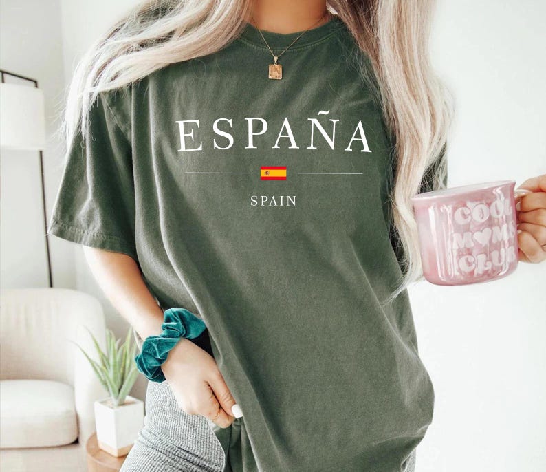 May include: Olive green t-shirt with the word "ESPA&Ntilde;A" printed in white, along with the Spanish flag and the word "SPAIN" underneath. The person is wearing a teal scrunchie and holding a pink mug.