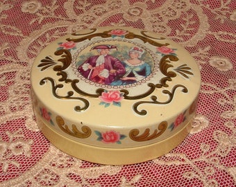 May include: A round, cream-coloured tin box with a gold-coloured design. The lid features a scene of two people in 18th-century clothing, surrounded by gold-coloured swirls and flowers.