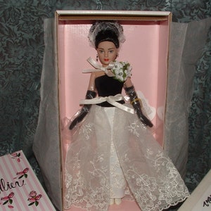 May include: A porcelain doll wearing a white lace dress with a black bodice and a bouquet of white flowers. The doll has black hair and brown eyes. The doll is in a pink box with white trim.