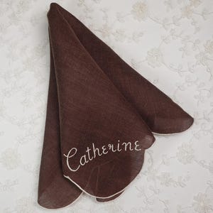 May include: A folded brown handkerchief with a white scalloped edge. The name "Catherine" is embroidered in white script on the front. The handkerchief is made of a lightweight fabric and is displayed against a white floral background.