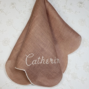 May include: A brown handkerchief with a scalloped edge and the name "Catherin" embroidered in white script. The fabric has a subtle woven texture. The handkerchief is folded, showcasing its shape and design.