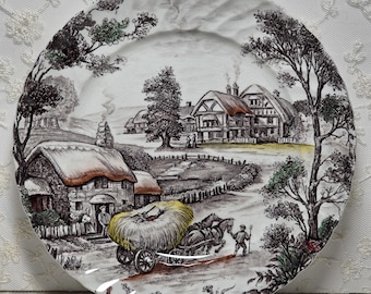May include: A decorative plate with a rural landscape scene. The plate features a black and white illustration of a village with thatched-roof houses, trees, and a horse-drawn cart carrying hay. The plate has a scalloped edge.