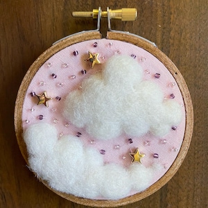 May include: A pink and white embroidery hoop with a fluffy white cloud and gold stars on a pink background. The hoop is decorated with small purple beads.