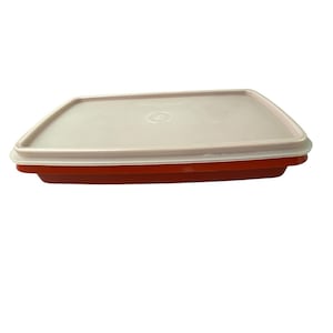 Puede incluir: Un recipiente rectangular para guardar alimentos con una base roja y una tapa rosa claro. La tapa tiene un borde ligeramente redondeado y una hendidura circular en el centro. El recipiente está diseñado para guardar alimentos.
