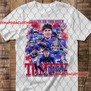 Puede incluir: Camiseta blanca con un gráfico de jugadores de fútbol americano en azul. El diseño incluye el texto "FIGHTING FOR YOUR DREAM" y "TOMMY DeVito" en rojo y azul. La camiseta tiene una superposición de valla de cadena.