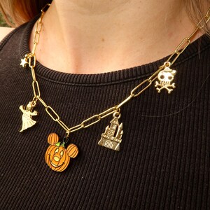 Può includere: Collana a catena color oro con ciondoli a tema Halloween. I ciondoli includono un fantasma, una stella, una zucca con la faccia di Mickey Mouse, un castello e un teschio. La collana è indossata su un top nero.