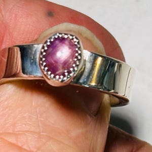 May include: A silver ring with a large, oval pink gemstone. The gemstone has a star-like effect and is set in a bezel with a beaded edge. The ring's band is wide and polished, reflecting light. The ring is a piece of jewellery.