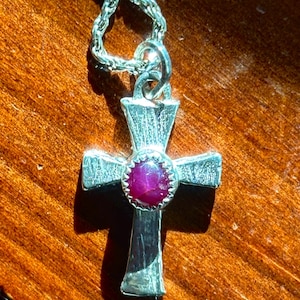 Peut inclure: Pendentif en forme de croix argenté avec une finition texturée. La croix présente une pierre précieuse rose foncé et ronde en son centre. La chaîne est de conception délicate et maillée. Le collier repose sur une surface en bois.