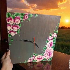 Może przedstawiać: Kwadratowy szary zegar ścienny z ręcznie malowanymi różowymi różami i zielonymi liśćmi. Zegar ma czarne wskazówki i pomarańczową wskazówkę sekundową. Zegar jest trzymany na tle zachodu słońca.