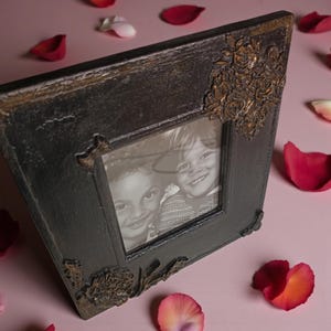 Puede incluir: Un marco de fotos negro con detalles florales en color bronce. El marco contiene una fotografía en blanco y negro de dos niños sonriendo. Pétalos de rosa están esparcidos sobre una superficie rosa.