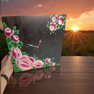 Może przedstawiać: Czarny kwadratowy zegar ścienny z kwiatowym wzorem. Zegar ma różowe i fioletowe róże z zielonymi liśćmi namalowanymi na rogach. Wskazówki zegara są srebrne i czerwone.
