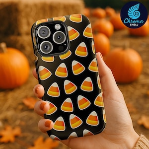 Puede incluir: Una funda de teléfono negra con un patrón repetido de caramelos de maíz. El caramelo de maíz es blanco, amarillo y naranja. La funda del teléfono se sostiene en una mano, con calabazas y balas de heno en el fondo.