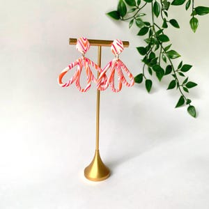 Puede incluir: Un par de pendientes colgantes con un patrón de remolinos jaspeados en blanco, rosa y naranja. Cada pendiente consta de un pequeño perno circular conectado a una pieza colgante más grande en forma de abanico abierto, sobre un soporte de metal dorado.