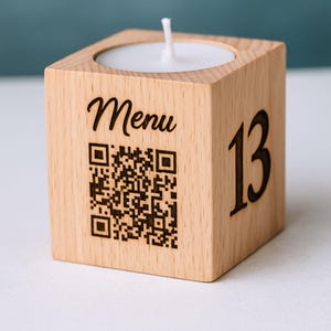 Può includere: Un portacandele in legno con una candela bianca. Il supporto a forma di cubo ha la parola "Menu" e un codice QR incisi su un lato. Il numero "13" è inciso su un altro lato.