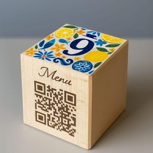 Pode incluir: Cubo de madeira clara com um topo decorativo com um grande número 9 azul, limões e padrões florais. O lado exibe a palavra "Menu" e um código QR.
