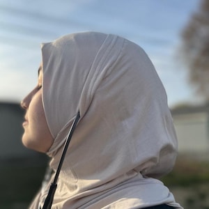 Può includere: Un hijab beige chiaro, un velo indossato da alcune donne musulmane. Il tessuto è drappeggiato morbidamente intorno alla testa e al collo. L'immagine è scattata con luce naturale, con uno sfondo sfocato di cielo blu e alberi.