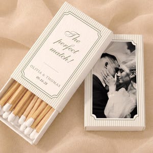 May include: Two rectangular matchboxes with white and beige pinstripes. One box is open, revealing wooden matches with white tips. The other box features a black and white photograph of a couple. Text reads: The perfect match! OLIVIA & THOMAS 05-16-28.