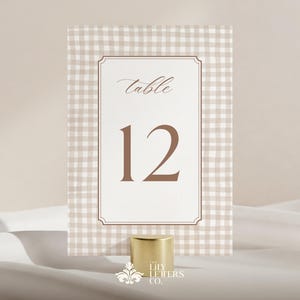 Op de afbeelding: Een tafelnummerkaart met een gingham patroonrand en een centraal paneel met het nummer 12 in bruin. Het woord "tafel" staat in script boven het nummer. Een goudkleurige basis ondersteunt de kaart.
