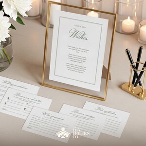 May include: A gold-framed sign with the words "Advice and Wishes" sits on a table, alongside pens in a gold holder and cards for guests to fill out. White peonies in a glass vase and candles complete the elegant wedding theme.