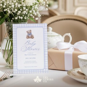 May include: A baby shower invitation featuring a teddy bear design, set against a blue and white gingham backdrop. The scene includes a gift box with a white ribbon, a teacup, and a vase of white flowers, all on a table.