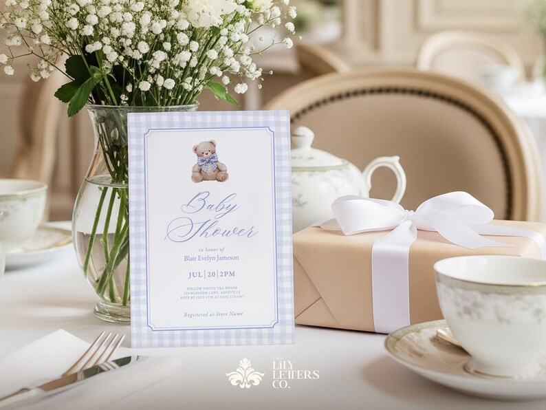 May include: A baby shower invitation featuring a teddy bear design, set against a blue and white gingham backdrop. The scene includes a gift box with a white ribbon, a teacup, and a vase of white flowers, all on a table.